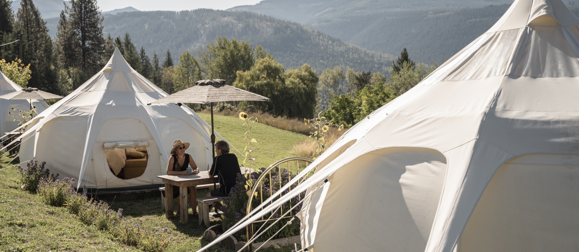 Two people sitting at a picnic table surrounded by mountains and glamping tents.
